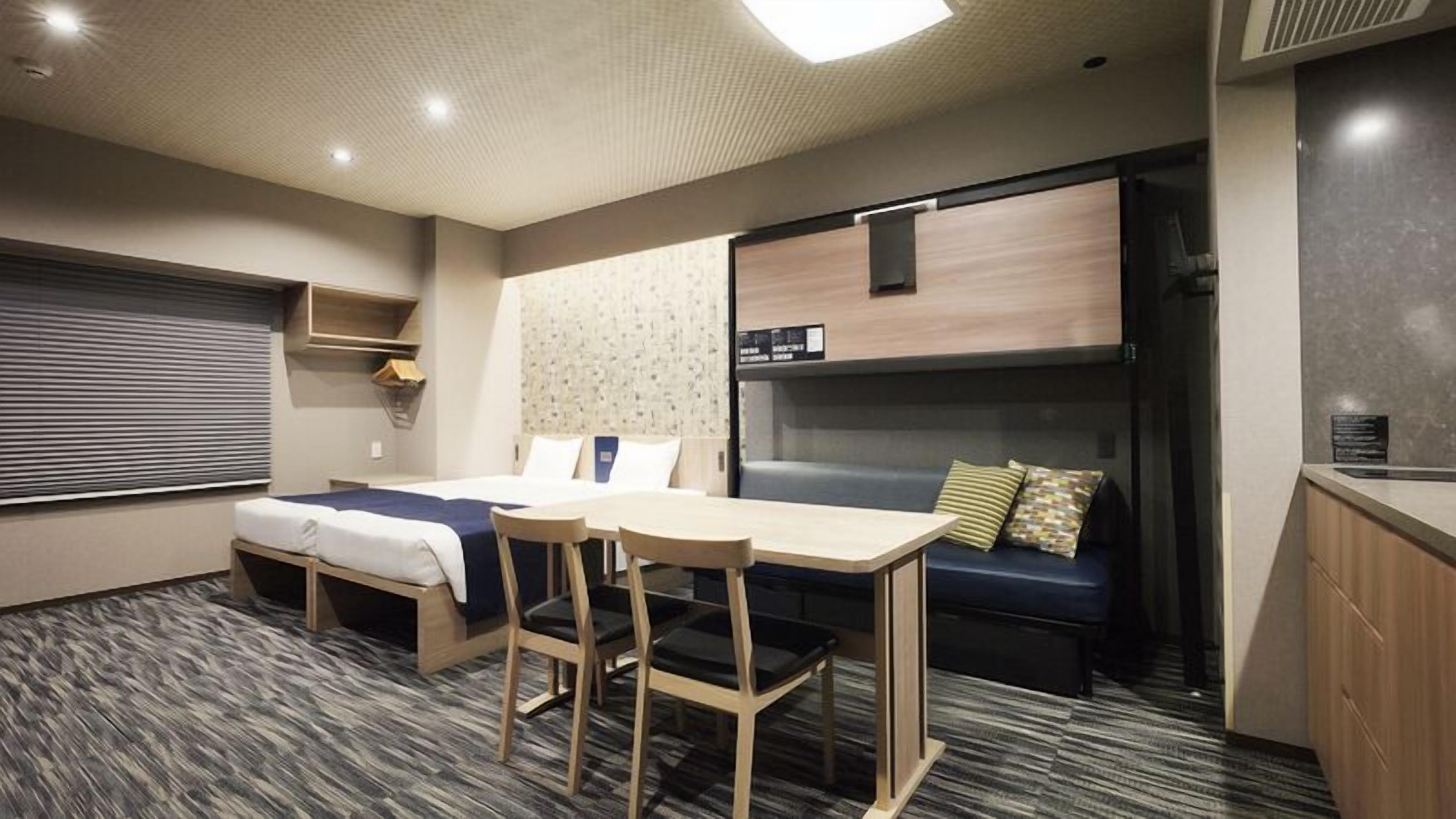 A compact hotel room with a bunk bed, a small table with chairs, a sofa, and a kitchenette.