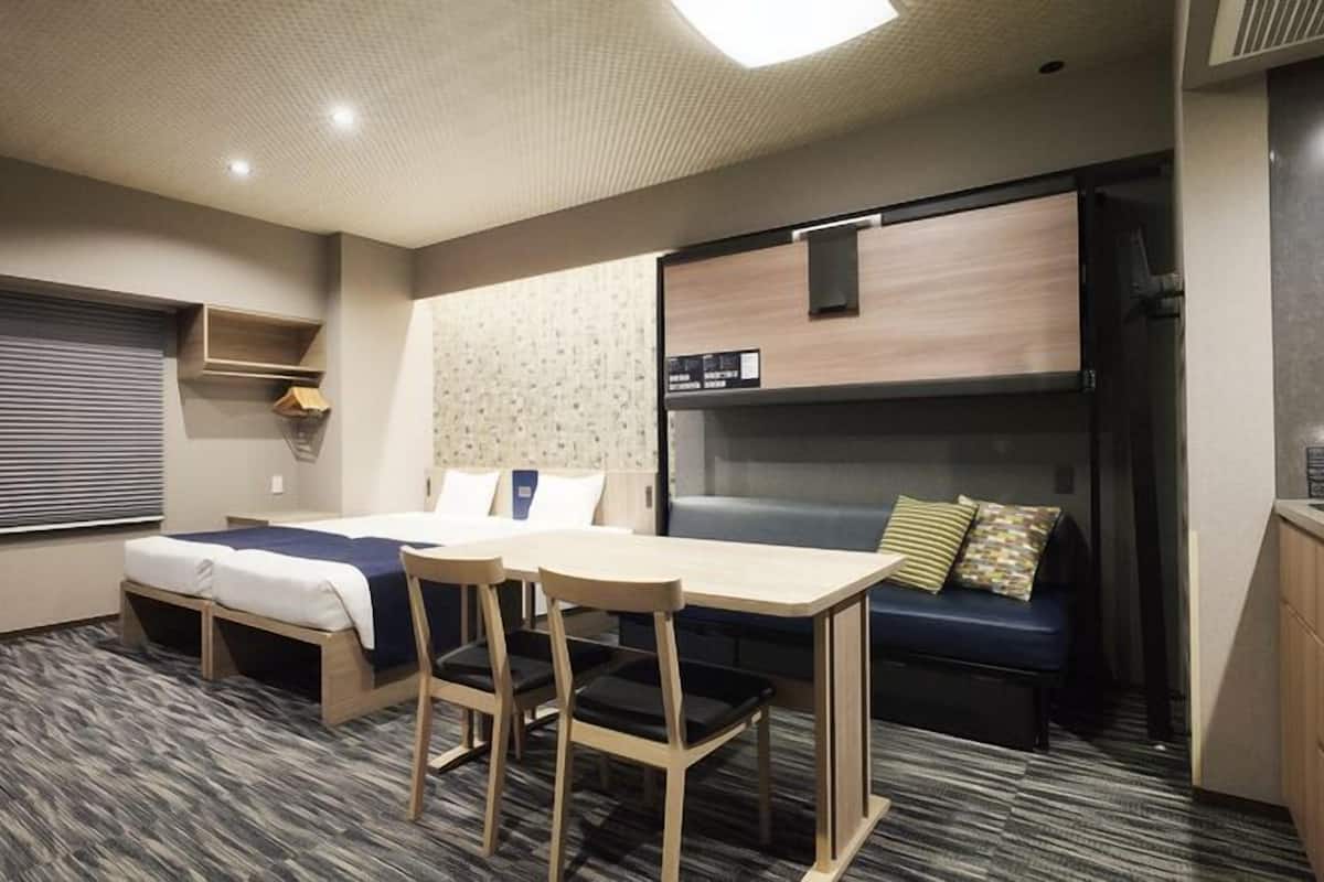A compact hotel room with a bunk bed, a small table with chairs, a sofa, and a kitchenette.