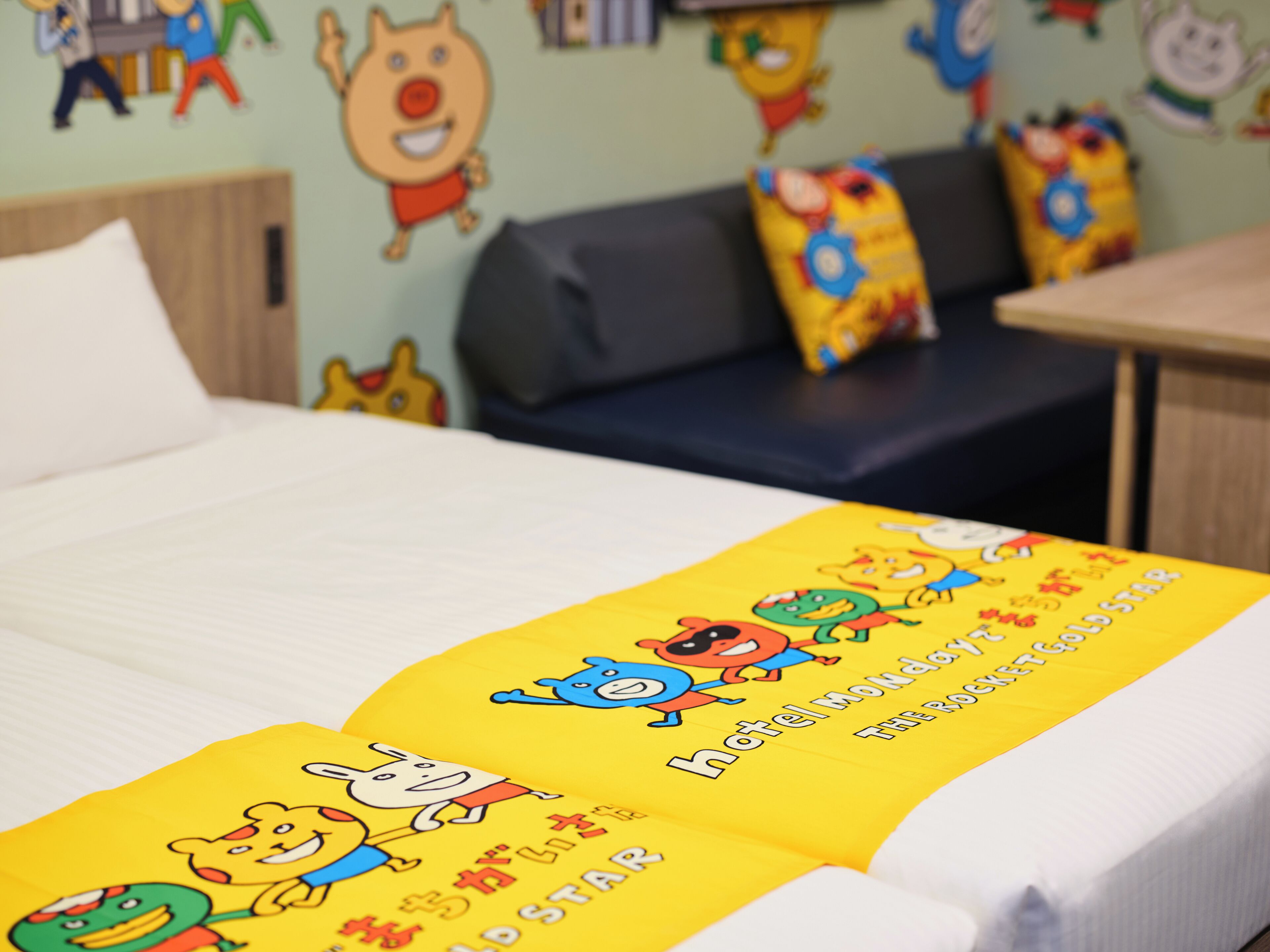 A hotel room with a bed, a sofa, and a table. The bed has a yellow blanket with cartoon characters.