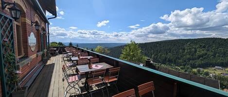 Una terraza con mesas y sillas de madera, un edificio de ladrillo con un letrero y vistas a un valle boscoso.