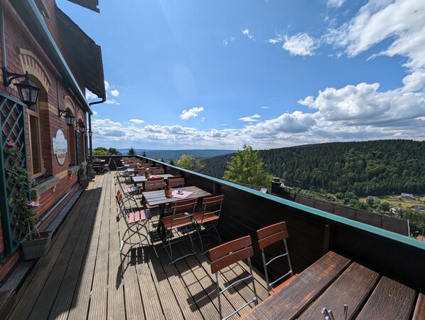 Eine Terrasse mit Holztischen und -stühlen, ein Backsteinhaus mit einem Schild und Blick auf ein bewaldetes Tal.
