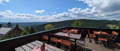 Una terraza de madera con mesas y sillas, con vista a un paisaje boscoso.