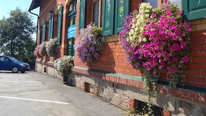 Un edificio de ladrillo con contraventanas verdes, un coche azul estacionado adelante y jardineras con flores en los laterales.