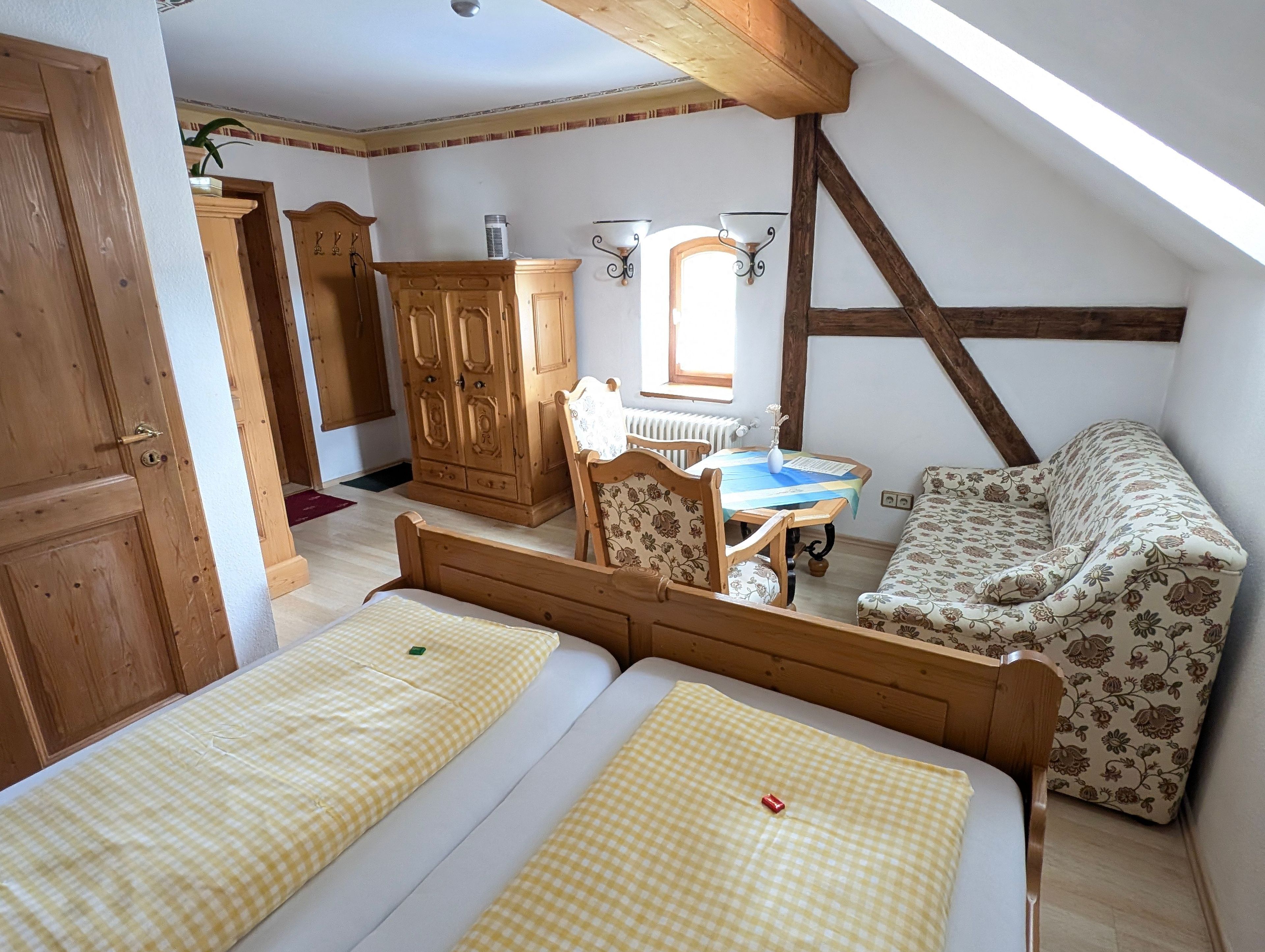 A cozy attic bedroom with two single beds, a wooden wardrobe, a floral sofa, and a small table with a vase.