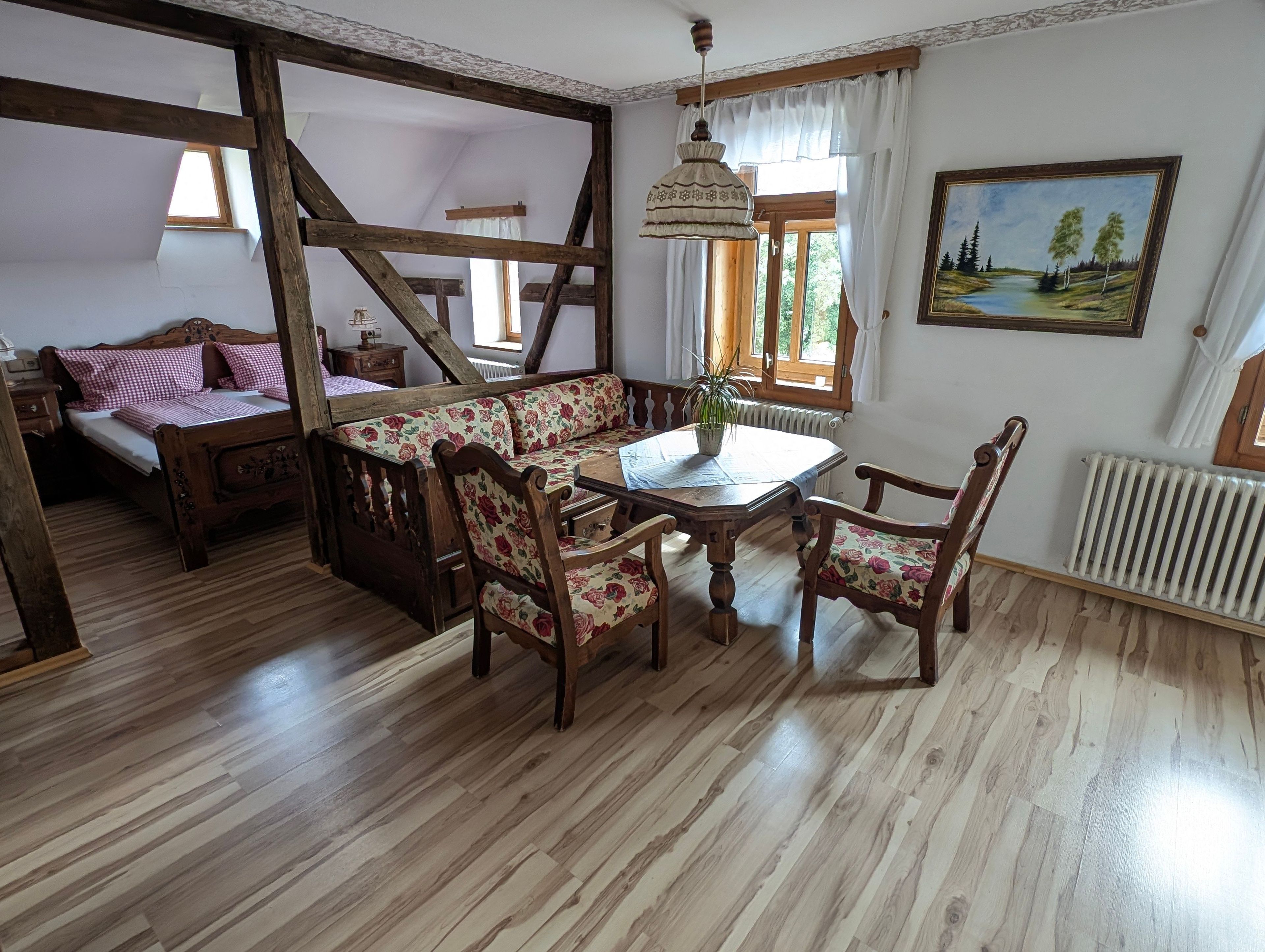 A room with a wooden floor, a bed with floral bedding, a wooden bench with floral cushions, a table with chairs, a hanging lamp, and a painting on the wall.