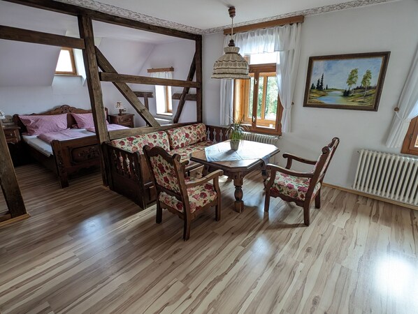A room with a wooden floor, a bed with floral bedding, a wooden bench with floral cushions, a table with chairs, a hanging lamp, and a painting on the wall.