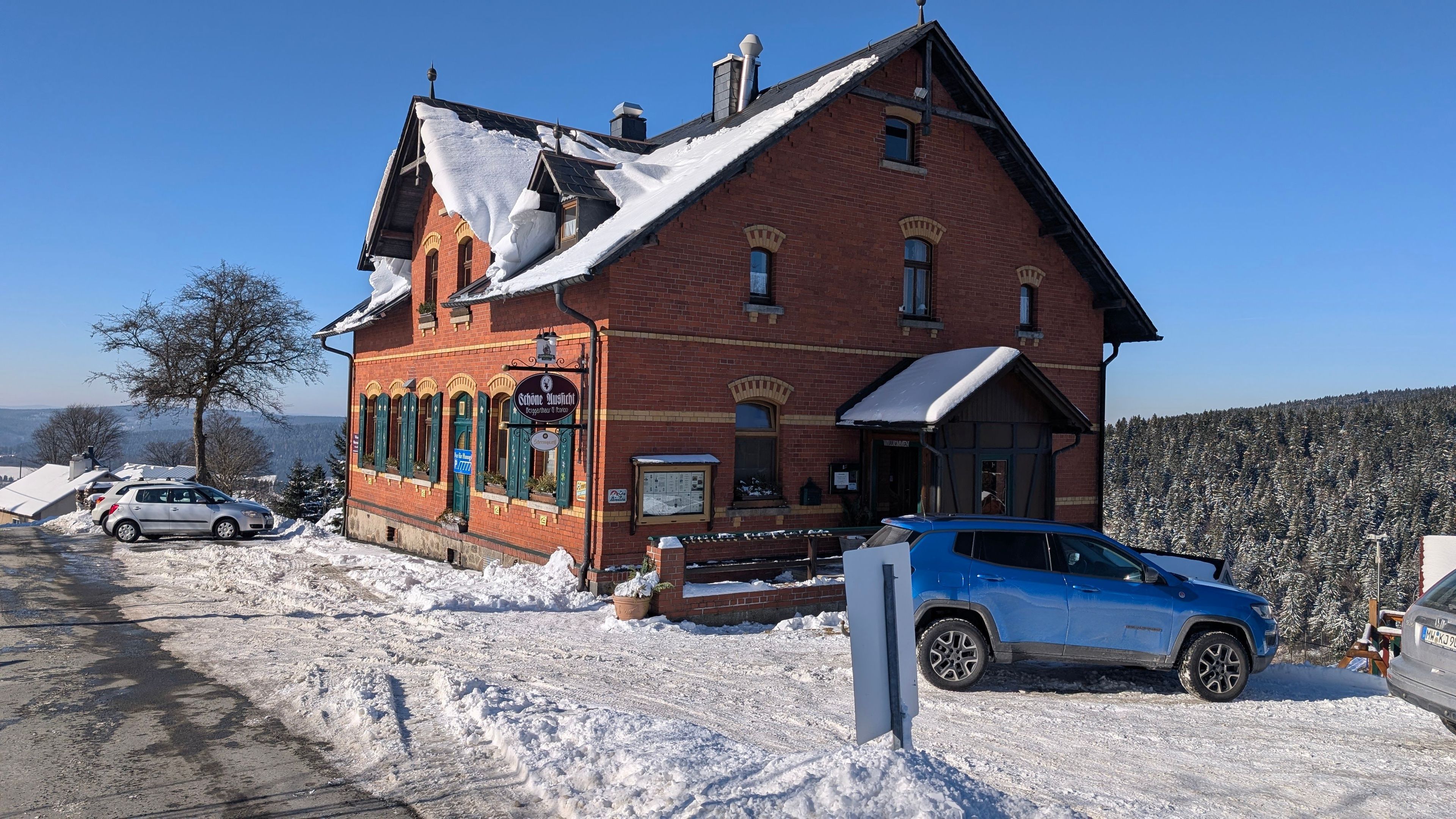 Un edificio de ladrillo rojo con techo cubierto de nieve, un letrero que dice 'Sahnehaus', y un coche azul estacionado.