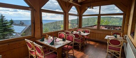 A wooden dining area with tables and chairs, large windows with scenic views, and a wooden ceiling.