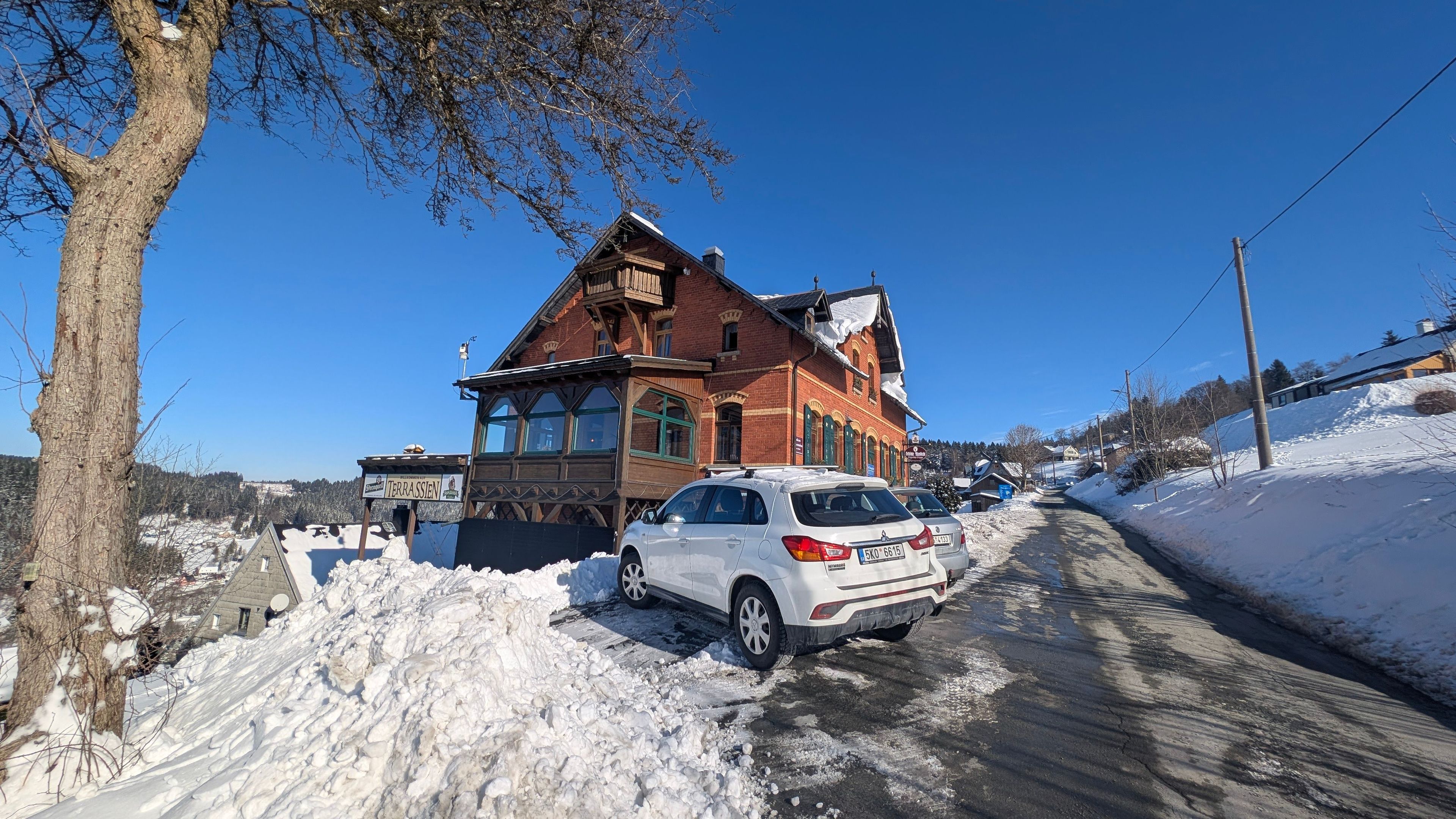 A snow-covered road with a white car parked in front of a traditional red brick building with a sign reading 'TERRASSEN'.