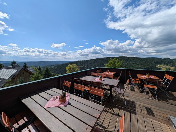 A wooden deck with tables and chairs, overlooking a forested landscape.