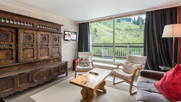 Un salon spacieux avec un meuble en bois, une table basse en bois, un canapé gris et une vue sur un paysage verdoyant grâce à de grandes fenêtres.