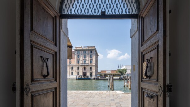A view through an arched doorway with wooden doors, leading to a canal with a building in the background.