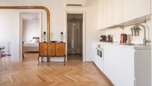 A modern kitchen with white cabinets, a wooden herringbone floor, and a small dining area with a wooden cabinet and a chair. - Venetian Palace Red Apartment R&R (Venecia)