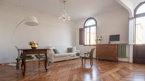 A spacious living room with a wooden herringbone floor, a white sofa, a dining table with chairs, a chandelier, and a large window with a view of buildings.