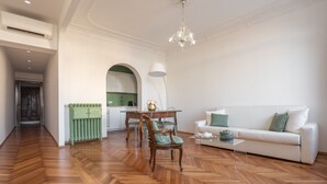 A spacious living room with a wooden floor, a white sofa, a dining table with chairs, and a kitchen in the background.