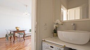 A bathroom with a large white sink, a mirror above it, and a toilet. There is a small table with a vase of flowers and a green chair in the adjacent room.