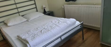 A neatly made bed with white linens in a room with wooden flooring, a radiator, and a window with a plant on the sill.