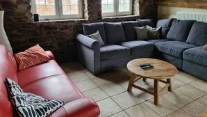 A cozy living room with a stone wall, two windows, a red leather sofa, a grey sectional sofa, and a wooden coffee table. - Beautiful very quiet Gite (Büllingen)