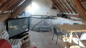 An attic room with a TV, chairs, a dining table, and a kitchen area with a stove and sink. - Beautiful very quiet Gite (Büllingen)