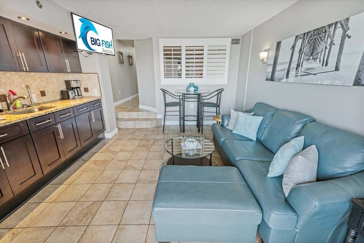A modern kitchen with dark cabinets, a granite countertop, and a coffee maker. There is a dining area with a table and chairs, a blue sofa, and a painting on the wall.