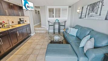 A modern kitchen with dark cabinets, a granite countertop, and a coffee maker. There is a dining area with a table and chairs, a blue sofa, and a painting on the wall.