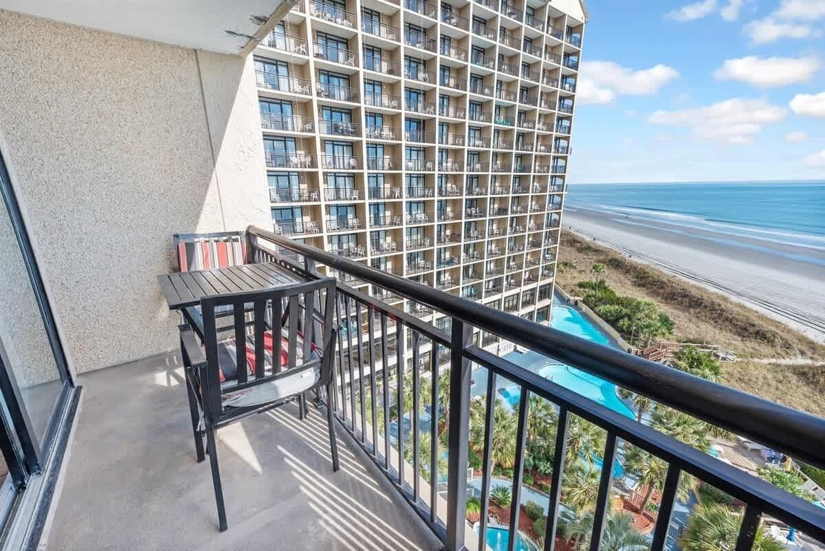 A balcony with a table and chairs, overlooking a beach and a hotel building with multiple balconies.