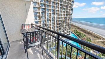 A balcony with a table and chairs, overlooking a beach and a hotel building with multiple balconies.