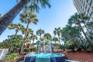 A pool with a fountain surrounded by palm trees and a building in the background.