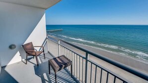 Ein Balkon mit zwei Korbstühlen und einem kleinen Tisch, der auf einen Strand mit ruhigen Wellen und einem strahlend blauen Himmel blickt.