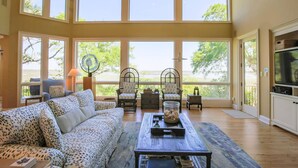 A spacious living room with large windows, a sofa, chairs, a coffee table, and a television.