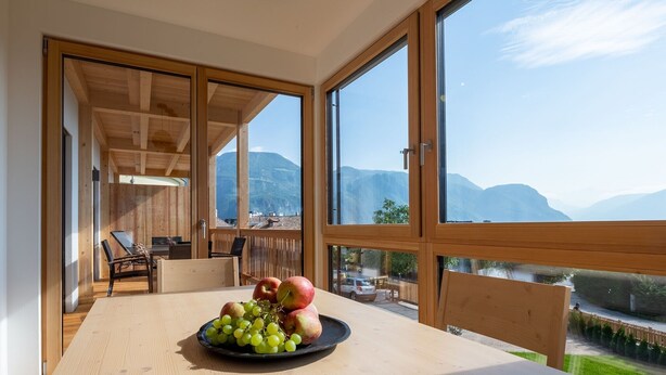 A room with a wooden table, a plate of fruit, and large windows with a mountain view. - Welcoming Apartment "Eiche im Nockerhof" with Mountain View, Wi-Fi & Terrace (Andrian)