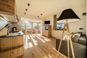 A modern kitchen with wooden cabinets, a dining area with a table and chairs, a sofa, and a large window with a view of mountains.