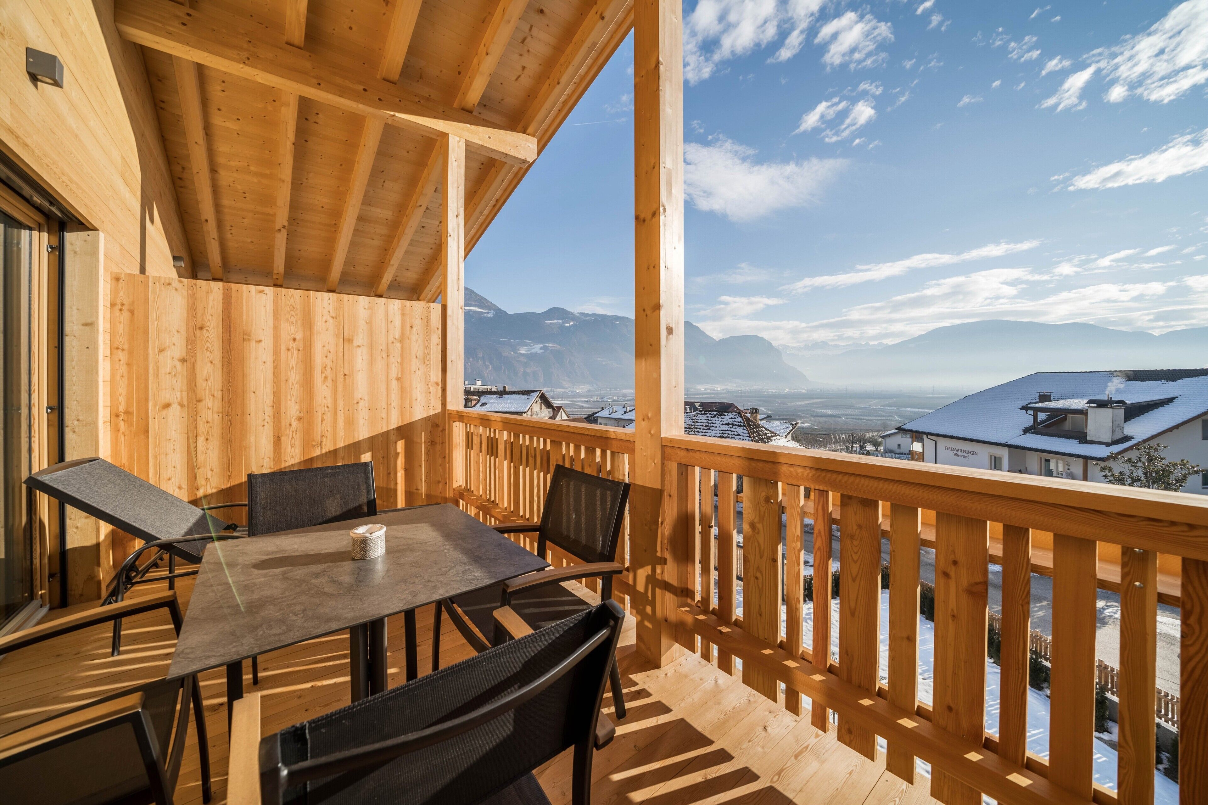 Un balcone in legno con tavolo e sedie, affacciato su un paesaggio innevato con edifici e montagne.