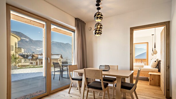 Un interno moderno con un tavolo da pranzo in legno, sedie, una lampada a sospensione e vista sulle montagne attraverso ampie porte scorrevoli.