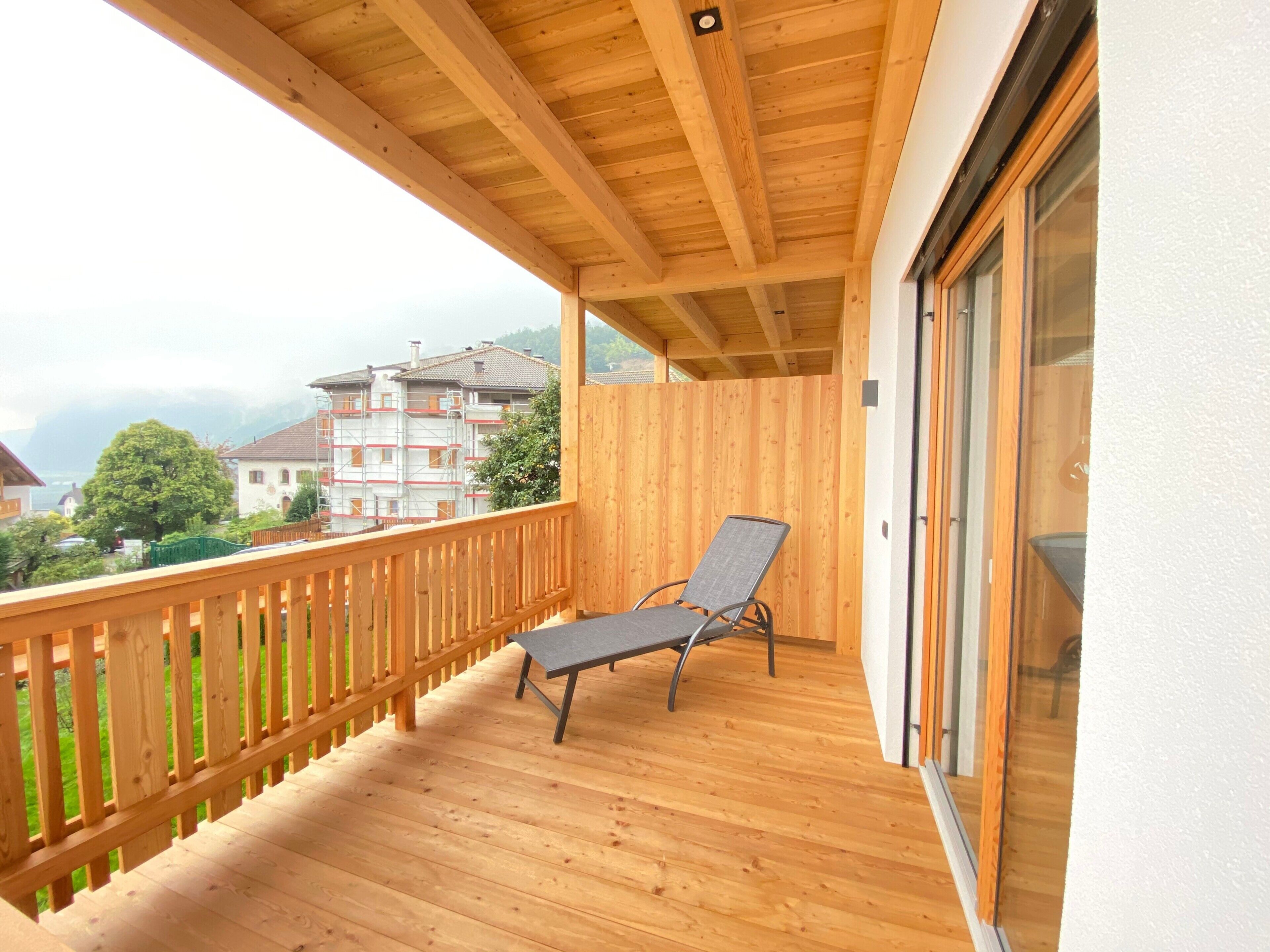 Un balcone in legno con una singola sedia, una ringhiera di legno e vista su edifici e alberi.