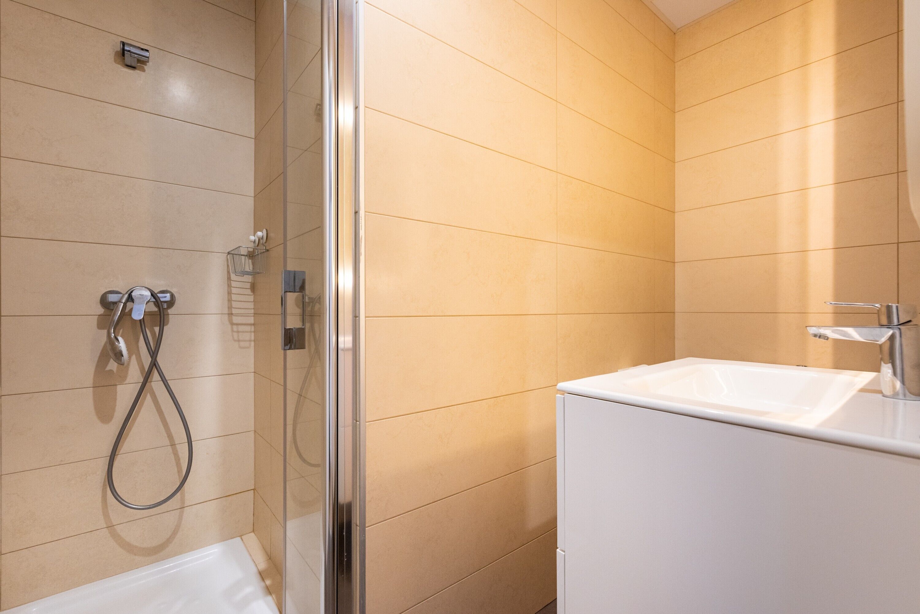 Une salle de bain moderne avec une douche à parois de verre, un lavabo blanc et des murs recouverts de carreaux beiges.
