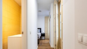 Un couloir étroit avec un plancher en bois, un lit recouvert d’une couvre-lit à motifs et une petite table avec une lampe.