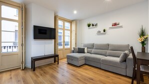 Un salon avec un canapé d’angle gris, un téléviseur à écran plat fixé au mur, un plancher en bois et une fenêtre donnant sur un balcon.