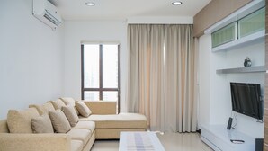 A modern living room with a beige sectional sofa, a white coffee table, a flat-screen TV mounted on the wall, and a large window with curtains. - Homey and Modern Tamansari Semanggi 2BR Apartment (Jakarta)