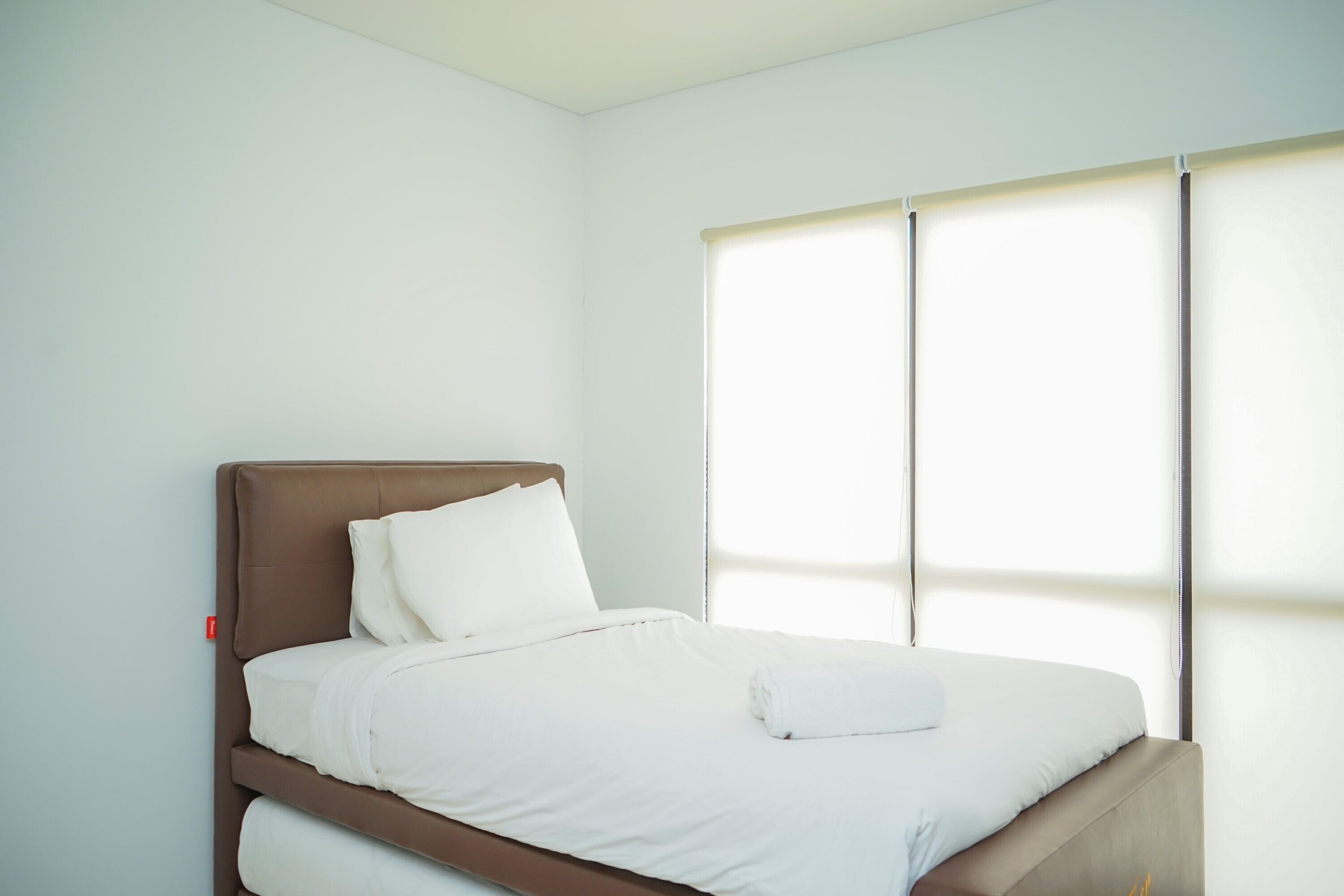 A neatly made bed with white linens and a brown headboard in a room with white walls and a large window with blinds.