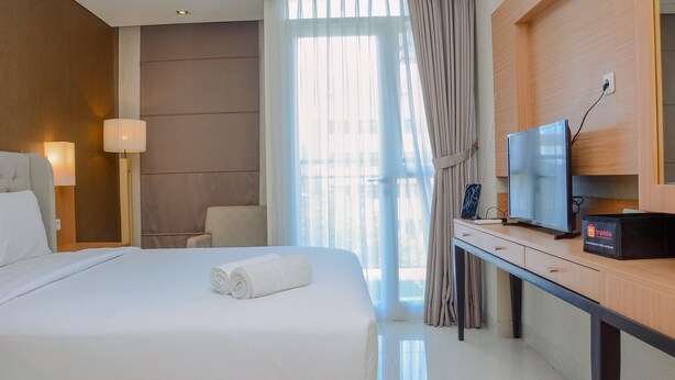 A hotel room with a large bed, a desk with a television, and a view of the city through the window. - Modern and Tidy Studio Apartment at Elpis Residence (Jakarta)