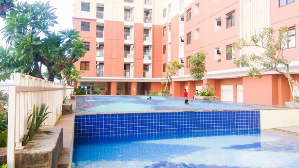Ein Swimmingpool mit blauen Fliesen, umgeben von einem weißen Zaun und viel Grün. Im Hintergrund ein mehrstöckiges Gebäude mit Balkonen und Fenstern.