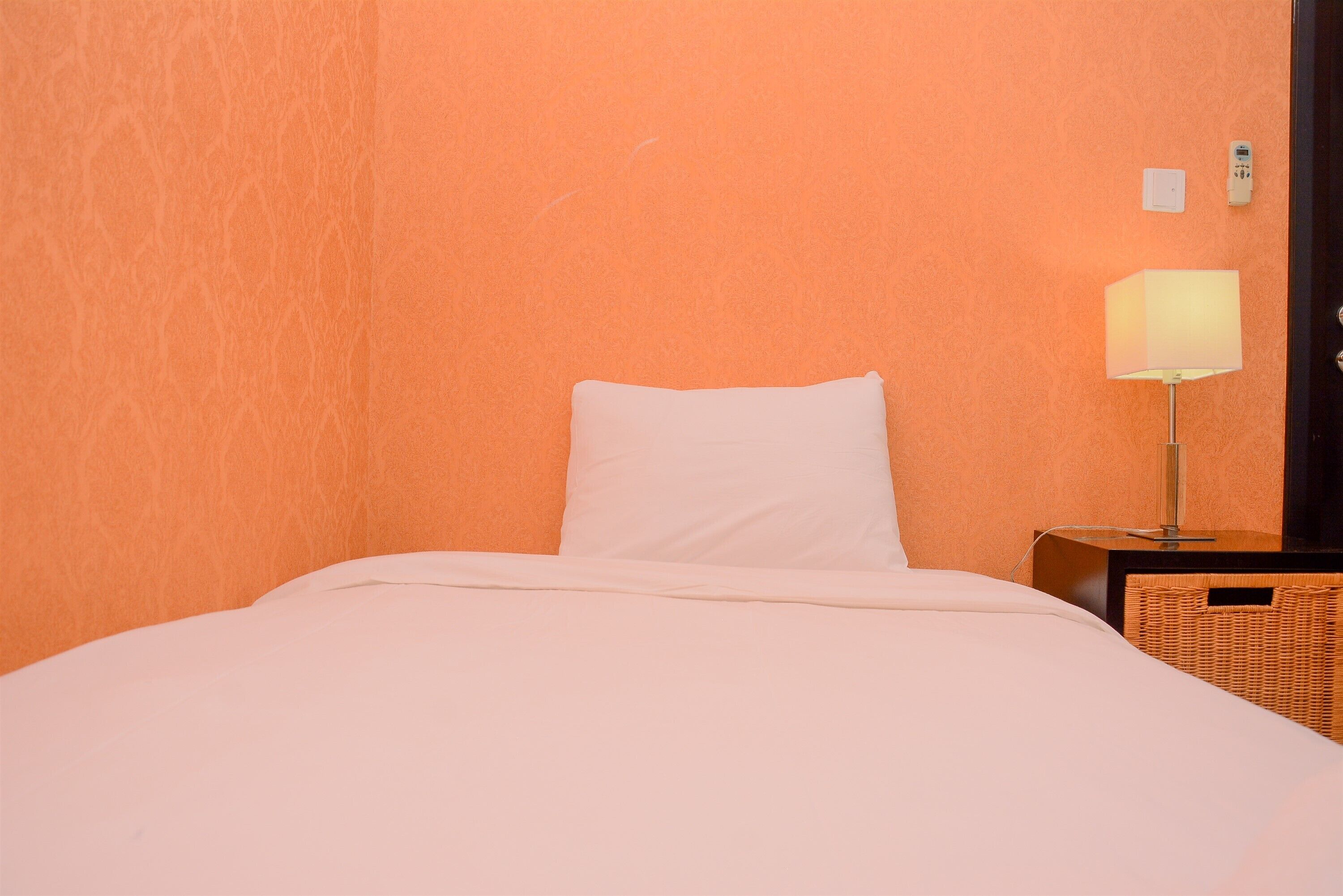 Uma cama bem arrumada com travesseiro e cobertor brancos, uma luminária de cabeceira e um criado-mudo de vime em frente a uma parede laranja.
