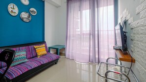 Una habitación con pared azul, un sofá con cojines de colores, una mesita, un televisor sobre una estantería de madera y una ventana con cortinas moradas.
