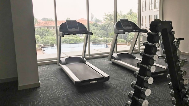 Un gimnasio con cintas de correr y mancuernas, amplias ventanas y vistas a edificios y árboles.