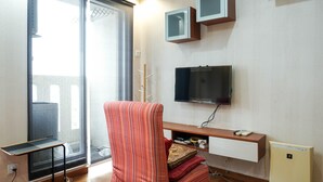 Een moderne woonkamer met een groot raam, een flatscreen televisie aan de muur, een kleine tafel en een bank met kussens.