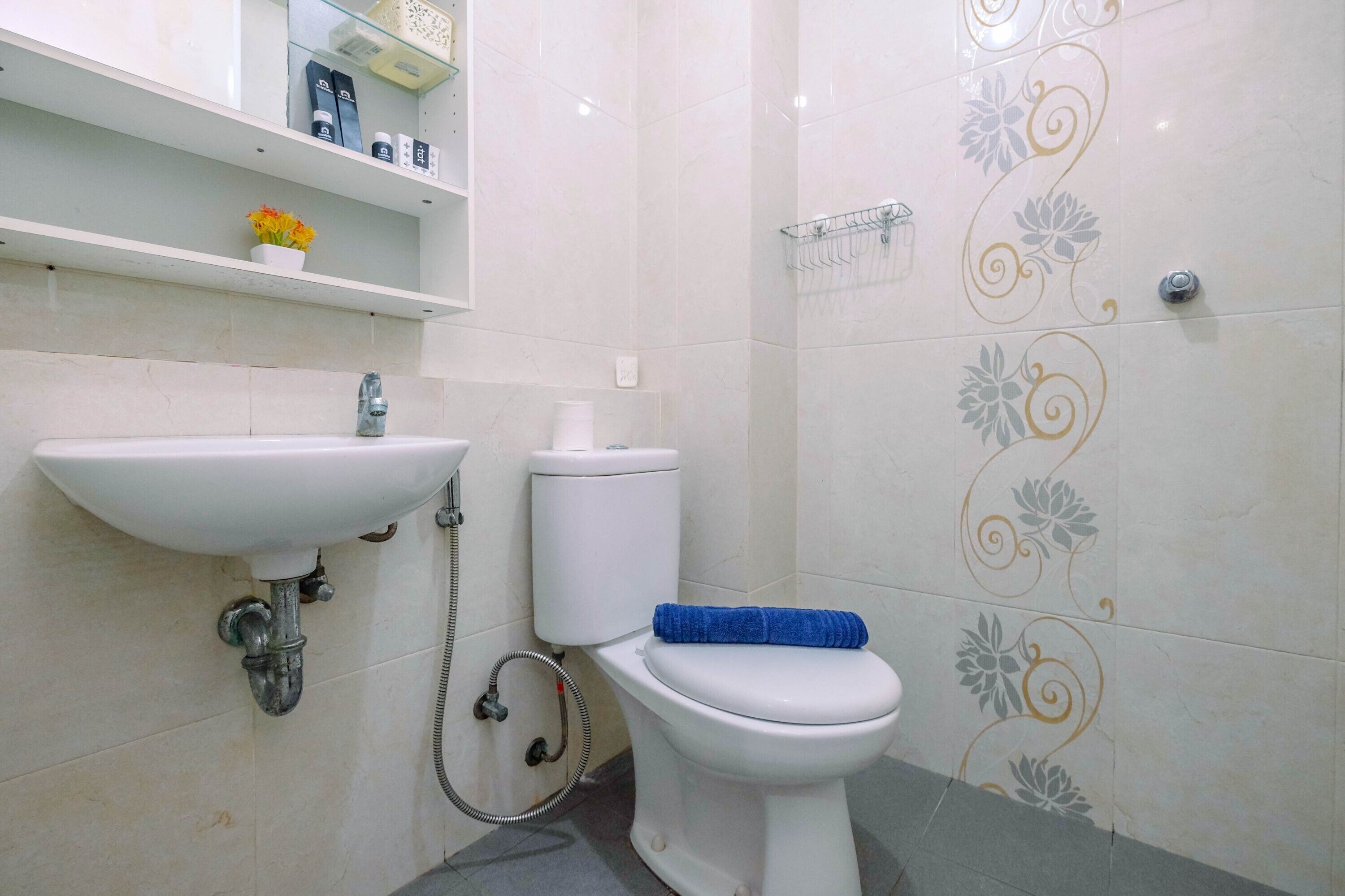 A bathroom with a white sink, toilet, and shower. There is a shelf with toiletries and a flower vase.
