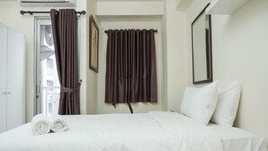 Habitación de hotel con una cama, ropa de cama blanca, dos cortinas marrones, una puerta blanca y un espejo en la pared.