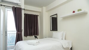 Habitación de hotel con cama, una puerta con cortinas, ventana con cortinas, estante montado en la pared con dos objetos, y una puerta que da a un balcón.