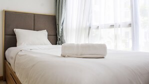 Un lit bien fait, avec une couette blanche et des oreillers, une tête de lit grise et une serviette blanche posée sur le lit.